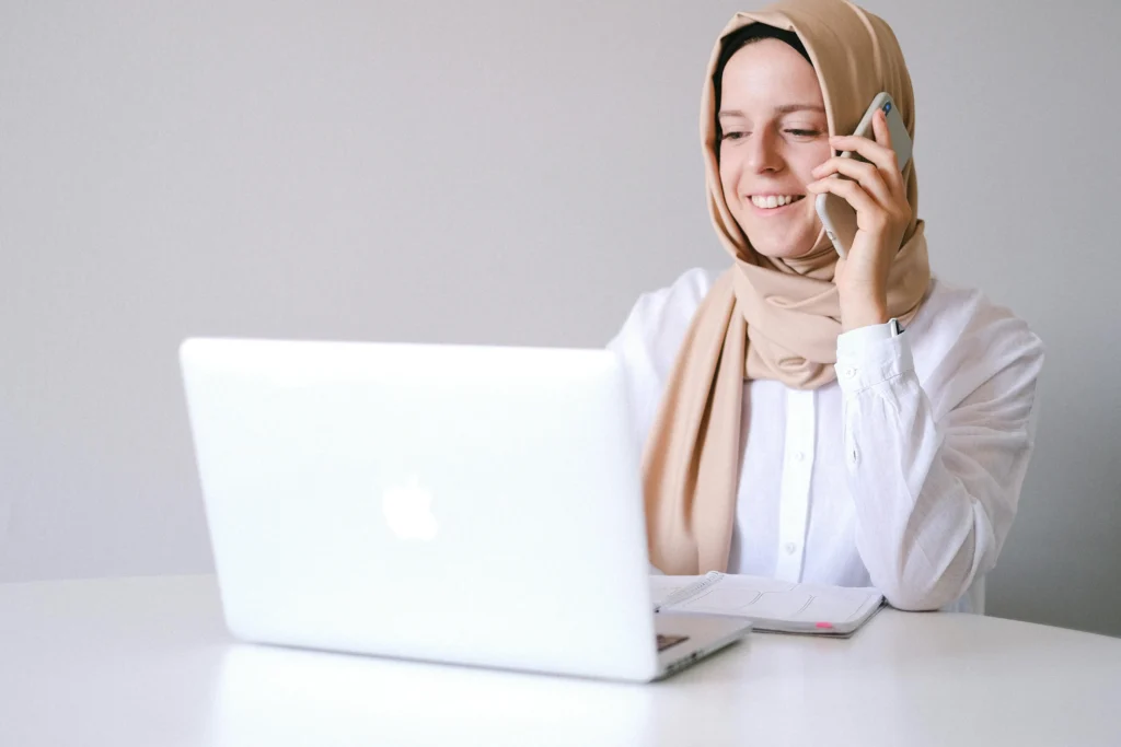 امرأة تجلس أمام حاسوب محمول وتمسك بهاتف ذكي بيدها، وتبحث عن أفكار لزيادة الدخل عبر الإنترنت.