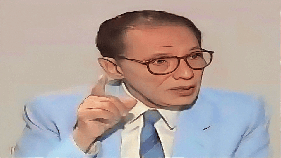 صورة للدكتور مصطفى محمود في مقابلة تلفزيونية يرتدي بزة رسمية وقبعة نظارات وهو يشير بإصبعه أثناء حديثه.