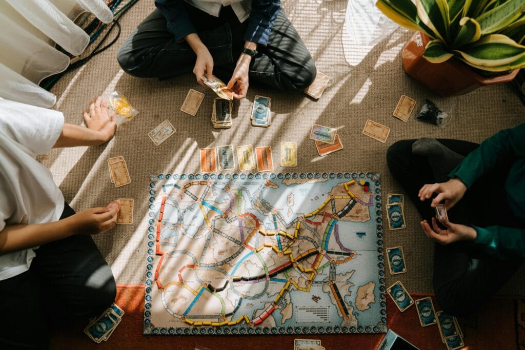 صورة لأطفال يلعبون بألعاب تعليمية مبتكرة في المنزل مع أحد الوالدين، تشمل مكعبات ملونة وألغازًا ولوحات تفاعلية لتنمية المهارات الحركية والعقلية