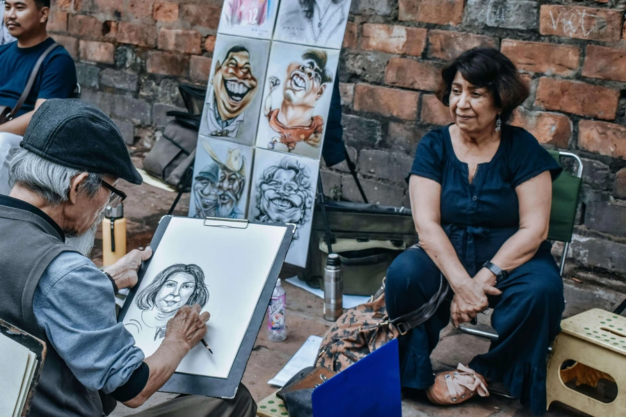 فنان شارع يرسم بورتريه واقعي لمرأة جالسة في الهواء الطلق مستخدمًا أقلام الرصاص على لوحة رسم محمولة.