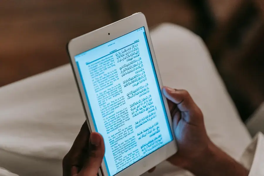 خص يقرأ كتاباً رقمياً على جهاز لوحي موضوع على طاولة، في بيئة مريحة تعكس التحول من الورق إلى الشاشة.
