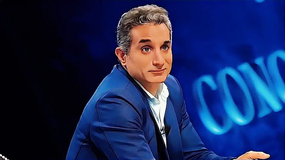 صورة للإعلامي والكوميدي باسم يوسف ينظر مباشرة إلى الكاميرا بتعبير وجهي معبر، يرتدي بدلة زرقاء وقميصًا أبيض، على خلفية استوديو يظهر جزء من كلمة "CON".