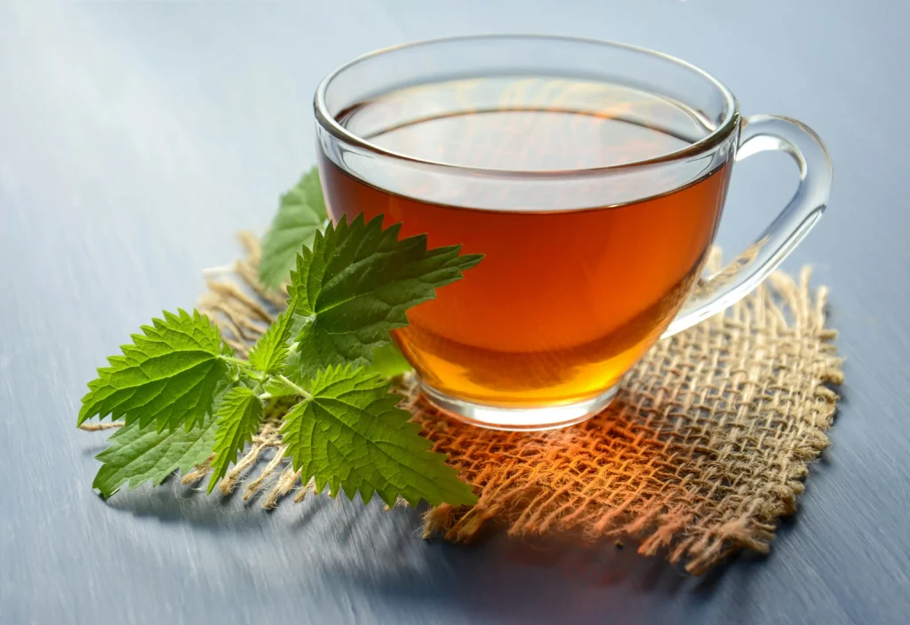 كوب شاي أعشاب دافئ موضوع على قطعة خيش طبيعية، بجانبه أوراق نباتية خضراء طازجة، يعبّر عن الراحة والعلاج الطبيعي.