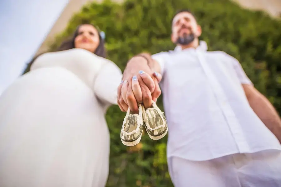 رجل وامرأة حامل يمسكان بأيدي بعضهما، بينما يحملان حذاء طفل صغير بين أصابعهما، في لقطة تُجسّد بداية رحلة الأمومة والأبوة.