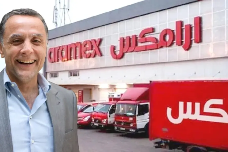فادي غندور مبتسم أمام مركز شركة أرامكس للشحن، وشاحنات حمراء تحمل شعار الشركة.