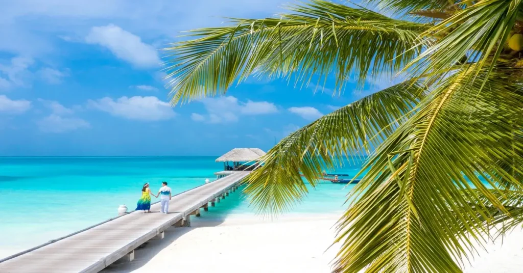 شاطئ أبيض ورصيف خشبي يمتد نحو البحر مع زوجين يستمتعان بجمال الطبيعة الساحلية.