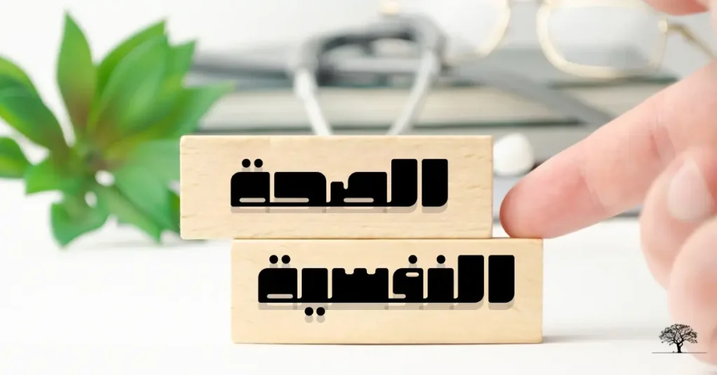 مكعبان خشبيان مرصوفان فوق بعضهما البعض، مكتوب عليهما كلمة "الصحة" و"النفسية" باللغة العربية، بينما يشير إصبع إلى الكلمة السفلية، يرمز إلى التركيز على أهمية التوازن العاطفي والعناية بالنفس.