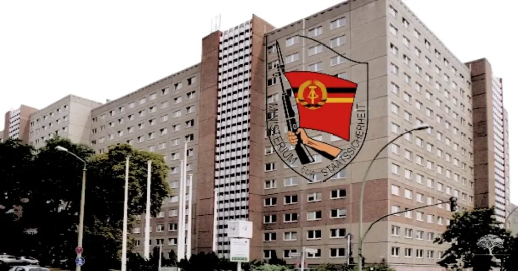 مبنى حكومي ضخم وممل الطراز، يمثل المقر الرئيسي لـ "جهاز شتازي" (وزارة أمن الدولة في ألمانيا الشرقية)، مع شعار الجهاز مُركباً على الواجهة، يرمز إلى السيطرة والمراقبة الشاملة.